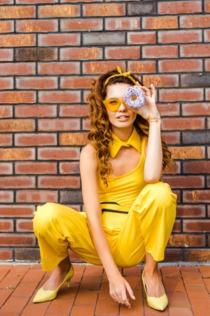 beautiful young woman in yellow clothes looking at camera through donut sitting in front of brick wallの写真素材
