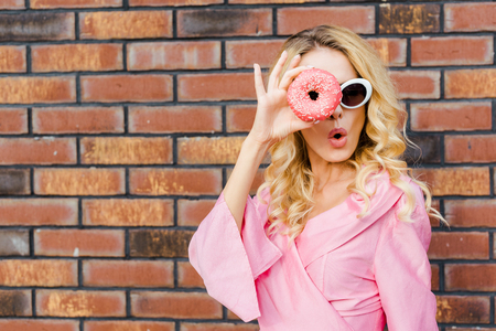 surprised young woman in pink shirt looking at camera through donut in front of brick wallの写真素材
