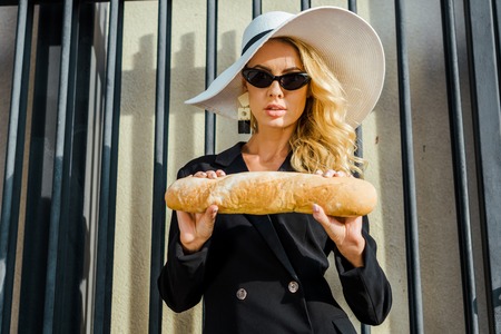 attractive young woman in stylish jacket and hat standing in front of fence with baguetteの写真素材