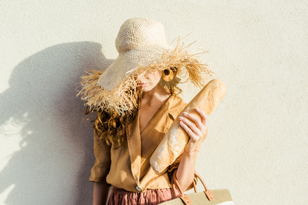 attractive young woman in stylish straw hat with baguette standing in front of white wallの写真素材