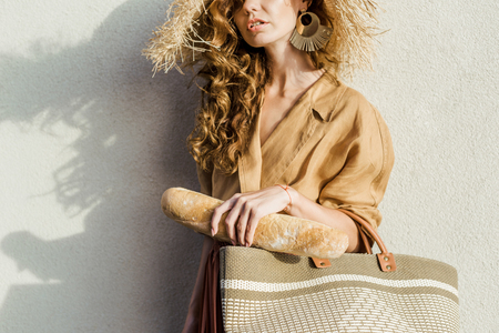 cropped shot of stylish woman in straw hat with baguette standing in front of white wallの写真素材