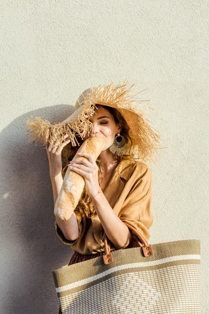 beautiful young woman in straw hat standing in front of white wall an eating baguetteの写真素材