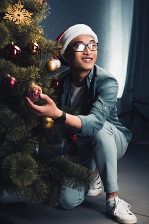 smiling asian man in santa hat decorating christmas tree and looking awayの写真素材