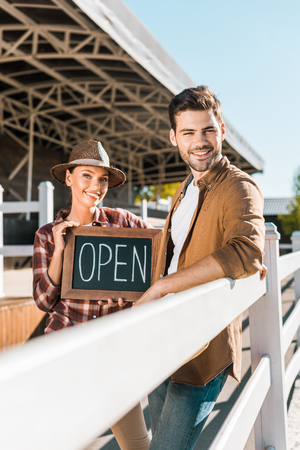 stylish cowboy and cowgirl in casual clothes standing near fence and holding open sign at ranchの写真素材