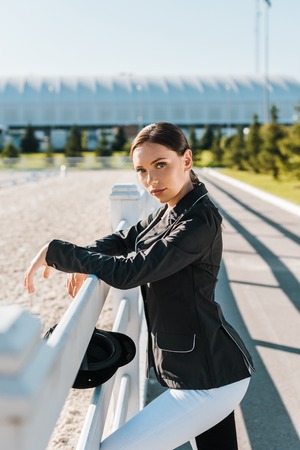 attractive female equestrian leaning on fence and looking at camera at horse clubの写真素材