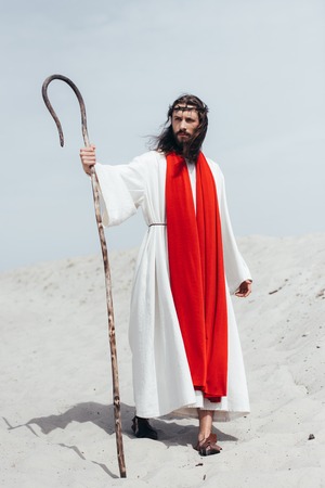 Jesus in robe, red sash and crown of thorns standing with wooden staff in desert and looking awayの写真素材