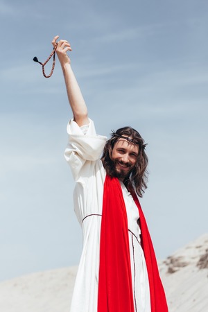 happy Jesus in robe, red sash and crown of thorns holding rosary in raised hand in desertの写真素材