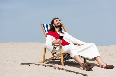 Jesus in robe and red sash resting on sun lounger with glass of red wine in desertの写真素材