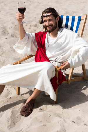 cheerful Jesus in robe and red sash resting on sun lounger and showing glass of red wine in desertの写真素材