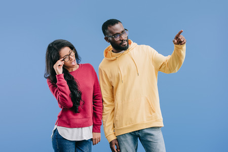 serious african american man pointing by finger to shocked girlfriend adjusting eyeglasses isolated on blue backgroundの写真素材