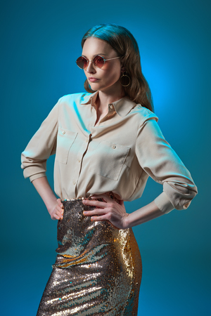 beautiful woman in fashionable golden glittering skirt and sunglasses posing with hands on waist isolated on blueの写真素材