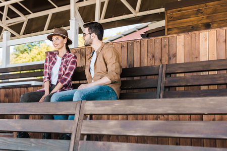 cheerful cowboy and cowgirl in casual clothes sitting on bench at ranch stadiumの写真素材