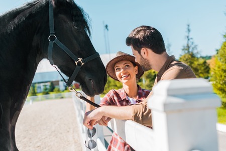 smiling cowboy and cowgirl standing near horse at ranch and looking at each otherの写真素材