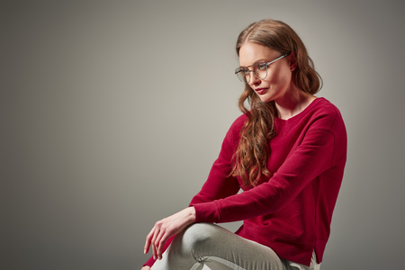 beautiful fashionable woman in eyeglasses sitting and looking away isolated on greyの写真素材