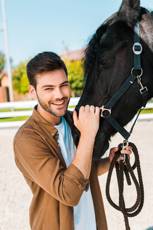 smiling handsome male equestrian holding horse halter and looking at camera at ranchの写真素材
