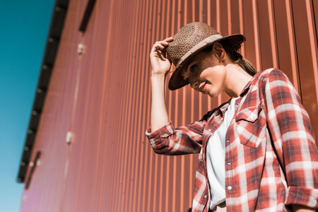 low angle view of attractive woman in checkered shirt and hat leaning on brown wall at ranchの写真素材