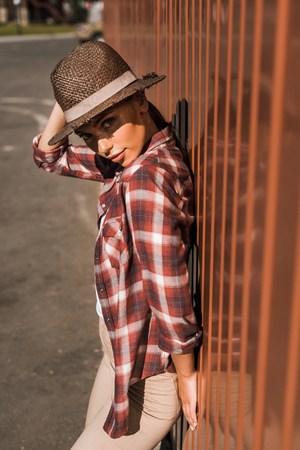 attractive woman in checkered shirt touching hat, leaning on brown wall at ranch and looking at cameraの写真素材