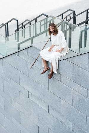 smiling Jesus in robe and crown of thorns sitting on staircase side, holding disposable coffee cup and looking downの写真素材