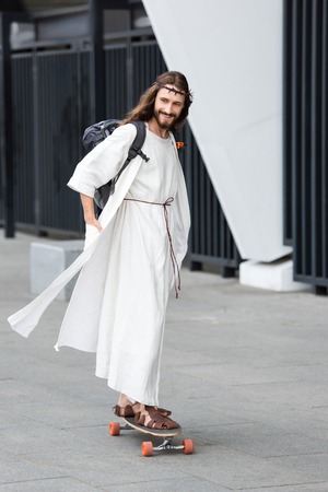 smiling Jesus in robe and crown of thorns skating on longboard on streetの写真素材