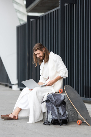 cheerful Jesus in robe and crown of thorns sitting on stone and using laptop on streetの写真素材
