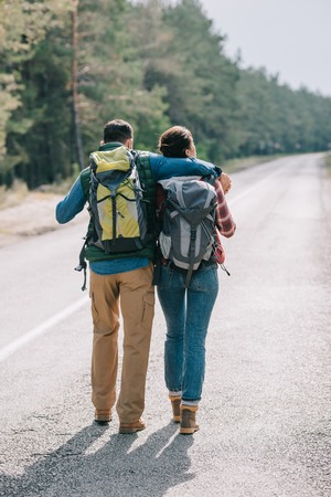 back view of couple of travelers with backpacks walking on roadの写真素材