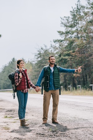 couple of travelers with backpacks holding hands hitchhiking on roadの写真素材