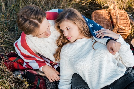 young couple with american flag resting on rural meadow, independence day concept