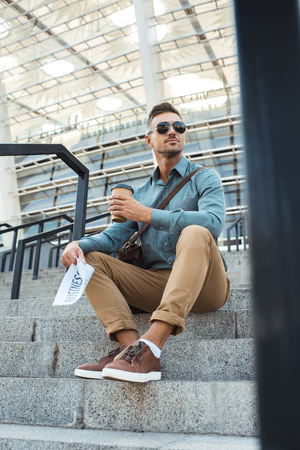 low angle view of man in sunglasses holding coffee to go and business newspaper while sitting on stairsの写真素材
