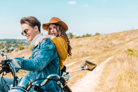 girlfriend looking at camera while boyfriend sitting on vintage motorbike on rural meadowの写真素材