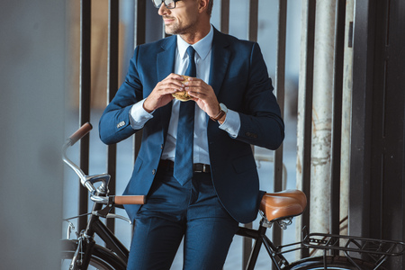 cropped shot of handsome middle aged businessman sitting on bike and holding hamburgerの写真素材