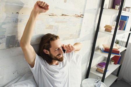 high angle view of bearded man with long hair doing stretch and talking on smartphone bed during morning time at homeの写真素材