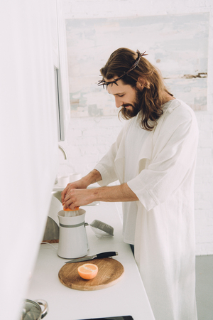side view of cheerful Jesus doing orange juice by juicer in kitchen at homeの写真素材