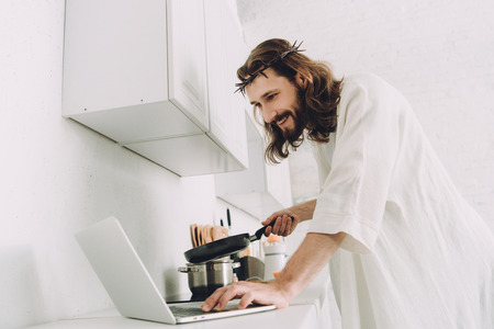 smiling Jesus using laptop while cooking with frying pan in kitchen at homeの写真素材