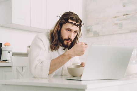 focused Jesus eating corn flakes on breakfast at table with laptop in kitchen at homeの写真素材