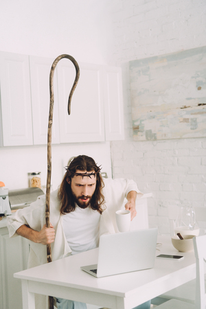 shocked Jesus with coffee cup and wooden staff looking at laptop in kitchen at homeの写真素材