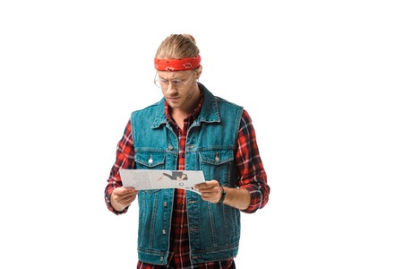 focused hipster man in denim vest reading travel newspaper isolated on whiteの写真素材