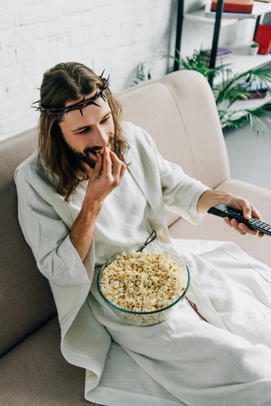 high angle view of Jesus in crown of thorns watching tv and eating popcorn on sofa at homeの写真素材