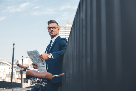 handsome buisnessman in suit and eyeglasses holding newspaper while standing with bike on streetの写真素材