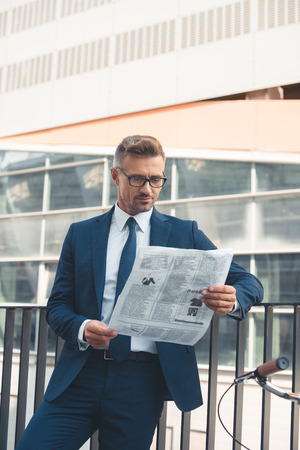handsome middle aged busnessman in suit and eyeglasses reading newspaper on streetの写真素材