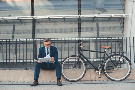 middle aged businessman reading newspaper while sitting near bicycle on streetの写真素材