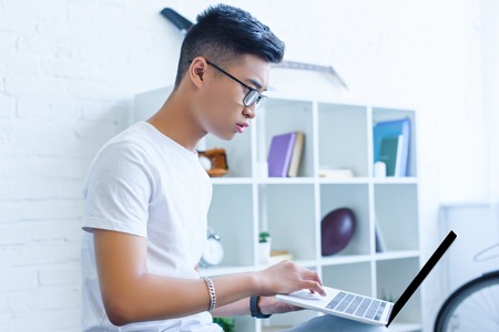 side view of handsome serious asian man using laptop on sofa at homeの写真素材