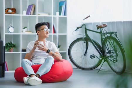 handsome asian man sitting on bean bag chair with cup of tea and looking away at homeの写真素材