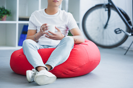 cropped image of man sitting on red bean bag chair with cup of tea in living roomの写真素材