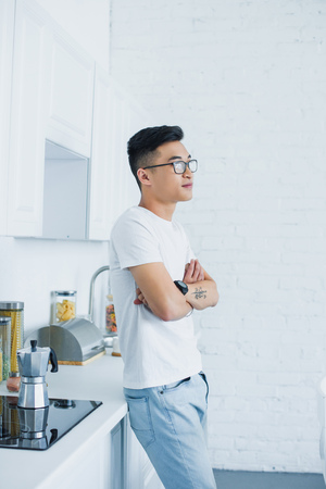 side view of handsome young asian man in eyeglasses standing with crossed arms and looking away in kitchenの写真素材