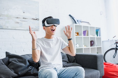 happy young asian man sitting on couch and using virtual reality headset at homeの写真素材