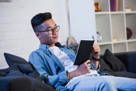 handsome smiling young man in eyeglasses sitting on couch and reading book