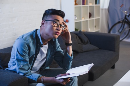 thoughtful young asian man in eyeglasses holding notebook and looking away at homeの写真素材