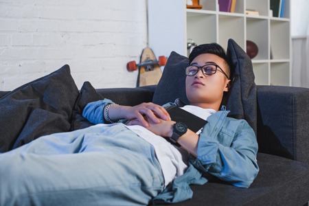 young asian man in eyeglasses holding book and sleeping on couchの写真素材