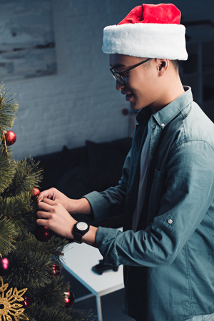 side view of smiling young asian man in santa hat decorating christmas tree at homeの写真素材
