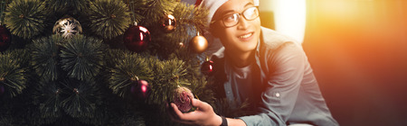 handsome happy young asian man in santa hat and eyeglasses decorating christmas tree and looking awayの写真素材